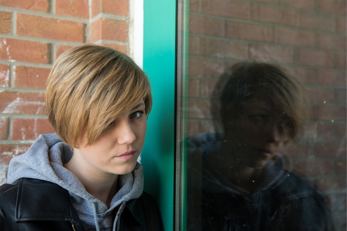 A young girl in a corner looking towards the camera, with a reflection in a window
